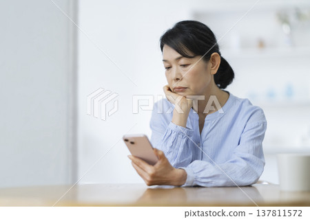 一位年近六十的女士一邊看著智慧型手機一邊沉思。 一位年近六十的女士一邊看著智慧型手機一邊沉思。 137811572