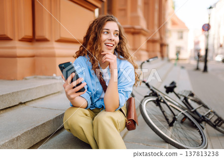 Woman using mobile phone, walks with bicycle, looks around at beautiful architecture. Selfie time. 137813238