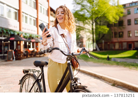 Woman using mobile phone, walks with bicycle, looks around at beautiful architecture. Selfie time. 137813251