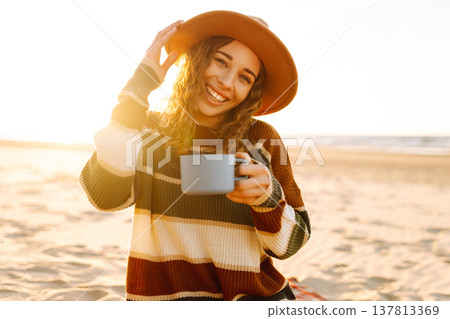 Happy woman in a stylish sweater sits at picnic on the beach drinks a hot drink from a thermos Happy woman in a stylish sweater sits at picnic on the beach drinks a hot drink from a thermos 137813369