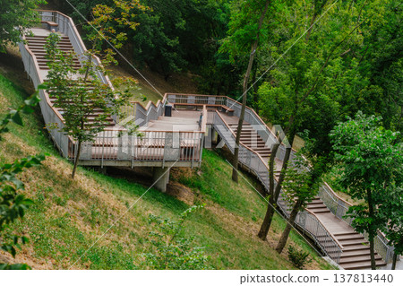 A high-angle horizontal shot of a long, zigzagging wooden staircase with metal railings built into a grassy hillside, surrounded by lush green trees in a park. A high-angle horizontal shot of a long, zigzagging wooden staircase with metal railings built into a grassy hillside, surrounded by lush green trees in a park. 137813440