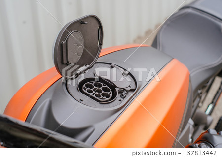 A high-angle horizontal shot of an orange electric motorcycle with the charging port cover open, showing the multi-pin connector on the fuel tank area, with the seat visible in the background. A high-angle horizontal shot of an orange electric motorcycle with the charging port cover open, showing the multi-pin connector on the fuel tank area, with the seat visible in the background. 137813442