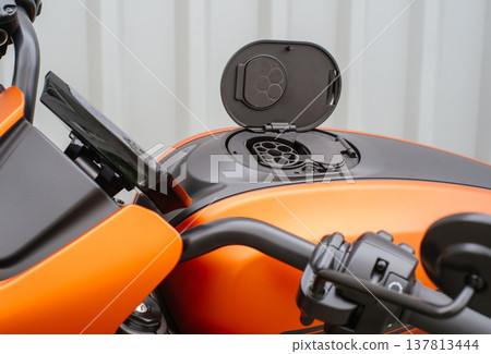 A horizontal shot from a side perspective showing the open charging socket on an orange electric motorcycle, with the handlebars and digital instrument cluster in the foreground against a neutral back 137813444