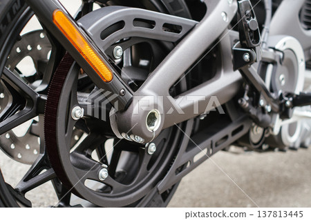 A detailed horizontal shot of the rear wheel assembly of a motorcycle, showcasing the belt drive system, brake disc, and a dark grey swingarm with an orange reflector. A detailed horizontal shot of the rear wheel assembly of a motorcycle, showcasing the belt drive system, brake disc, and a dark grey swingarm with an orange reflector. 137813445