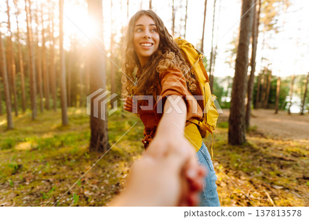 Follow me! Young woman joyfully exploring a sunny forest trail with a backpack, holding hands Follow me! Young woman joyfully exploring a sunny forest trail with a backpack, holding hands 137813578