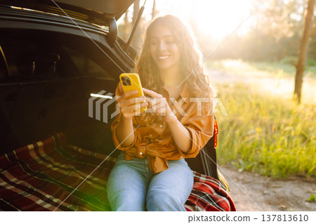 Young curly woman in stylish clothes in the trunk of a car in the forest and uses the phone. Young curly woman in stylish clothes in the trunk of a car in the forest and uses the phone. 137813610