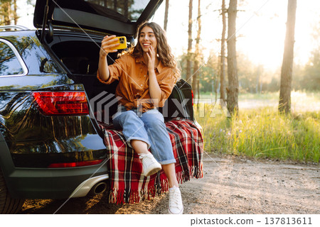 A joyful woman taking a selfie with her smartphone while sitting in the trunk of her car 137813611