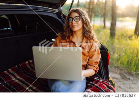 Happy woman working with laptop from the car trunk or boot in camper sunny summer park. 137813627