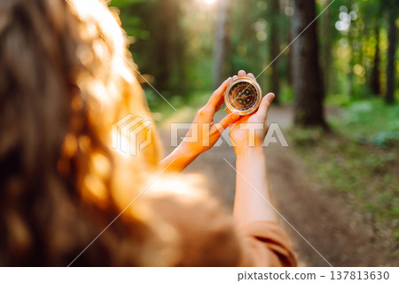 A hiker with curly hair holds a compass in their hands, carefully navigating through a sunlit forest 137813630