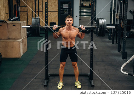 Exercise of squats with heavy barbell. Athletic man is in a gym Exercise of squats with heavy barbell. Athletic man is in a gym 137815016