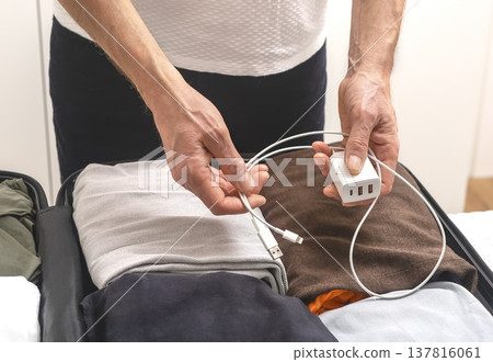 Charging cables and clothes inside open suitcase, Hands hold charger, wire above packed clothes in 137816061