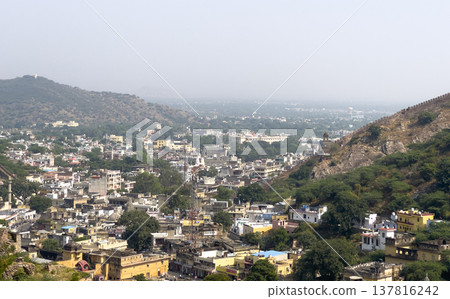 Jaipur cityscape fills valley beneath fortified hill and pale sky. Historic Rajasthan fort stands 137816242