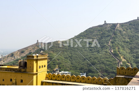 Jaipur fort wall climbs green hill ridge beneath pale sky. Yellow sandstone tower overlooks valley 137816271