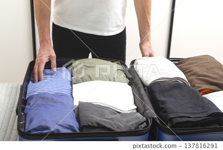 Man organizing neatly folded clothes inside blue suitcase. Male placing ordered garments in open luggage on bed. Travel planning, packing routine, departure readiness concept.. Man organizing neatly folded clothes inside blue suitcase. Male placing ordered garments in open luggage on bed. Travel planning, packing routine, departure readiness concept.. 137816291