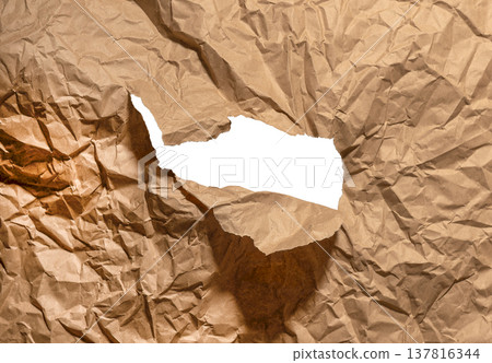 Ripped brown kraft paper forms irregular hole across crumpled textured surface. Jagged torn opening 137816344