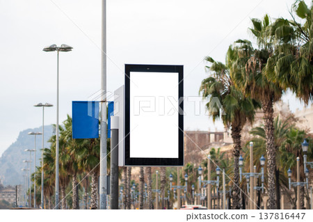 Blank vertical billboard on a city street, ready for custom advertising display, with urban architecture, green palm trees, and modern lampposts in a Mediterranean setting 137816447