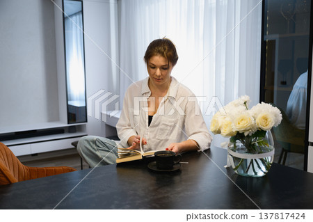 Young woman sitting at a table reading a book with a cup of coffee in a modern apartment. Young woman sitting at a table reading a book with a cup of coffee in a modern apartment. 137817424