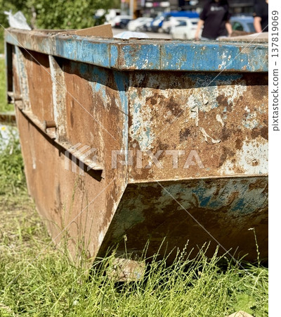 An Old and Rusty Dump Container That is Surrounded by Lush, Overgrown Grass and Weeds. A weathered metal dumpster overflowing with various debris, positioned against a backdrop of lush tall grass. 137819069