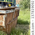 An Old and Rusty Dump Container That is Surrounded by Lush, Overgrown Grass and Weeds. A weathered metal dumpster overflowing with various debris, positioned against a backdrop of lush tall grass. 137819092