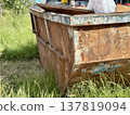 An Old and Rusty Dump Container That is Surrounded by Lush, Overgrown Grass and Weeds. A weathered metal dumpster overflowing with various debris, positioned against a backdrop of lush tall grass. 137819094