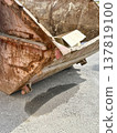 An old rusty dumpster sitting on a bustling city street filled with urban life and debris. The dumpster resting on the pavement clearly shows numerous signs of urban decay and waste management issues. 137819100