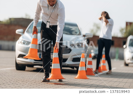 Placing the guide cones. Man instructor in the driving school and woman are outside the car Placing the guide cones. Man instructor in the driving school and woman are outside the car 137819943