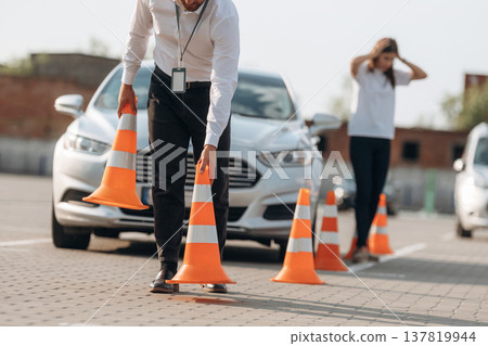 Placing the guide cones. Man instructor in the driving school and woman are outside the car Placing the guide cones. Man instructor in the driving school and woman are outside the car 137819944