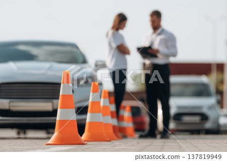 Talking with each other, cones on the road. Man instructor in the driving school and woman are outside the car Talking with each other, cones on the road. Man instructor in the driving school and woman are outside the car 137819949