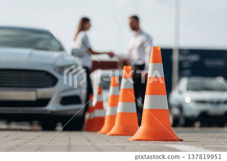 Talking with each other, cones on the road. Man instructor in the driving school and woman are outside the car Talking with each other, cones on the road. Man instructor in the driving school and woman are outside the car 137819951