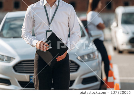Documents in hands of a guy. Man instructor in the driving school and woman are outside the car Documents in hands of a guy. Man instructor in the driving school and woman are outside the car 137820095