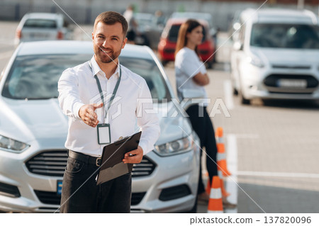 Documents in hands of a guy. Man instructor in the driving school and woman are outside the car Documents in hands of a guy. Man instructor in the driving school and woman are outside the car 137820096