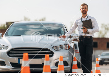 Leaning on the car. Driving school worker is outdoors Leaning on the car. Driving school worker is outdoors 137820195