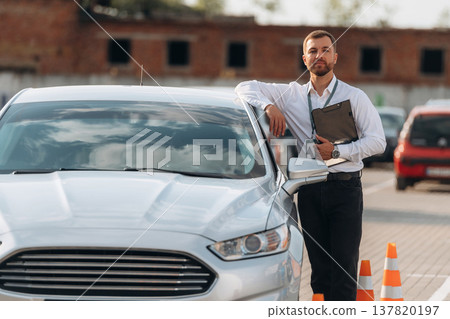 Leaning on the car. Driving school worker is outdoors Leaning on the car. Driving school worker is outdoors 137820197