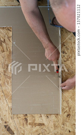 A utility knife in the hands of a repairman sitting on the floor and cutting a sheet of cardboard or plywood in a straight line, on the floor during repairs, top view, vertical image A utility knife in the hands of a repairman sitting on the floor and cutting a sheet of cardboard or plywood in a straight line, on the floor during repairs, top view, vertical image 137821112