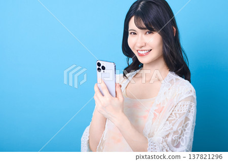 A smiling Japanese woman holding a smartphone against a blue background. (Copy space) A smiling Japanese woman holding a smartphone against a blue background. (Copy space) 137821296