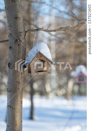 Snowy wooden birdhouse in nature. Rustic birdhouse covered in fresh snow. Peaceful winter scene showing rustic birdhouse adorned with fresh snow amidst textured bark background 137821605