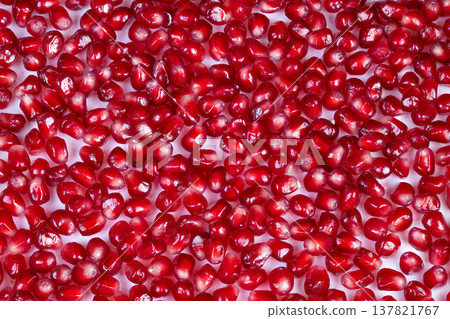 Shiny white surface hosts red seeds. Bright red arils scatter on glossy white background. Lustrous pomegranate seeds spread over smooth surface with vibrant color contrast 137821767