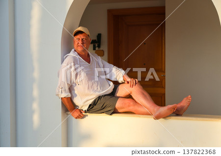 Adult man in a white shirt and cap sitting on a wide windowsill, enjoying the beautiful sunset, reflecting on life and tranquility in the golden hour 137822368