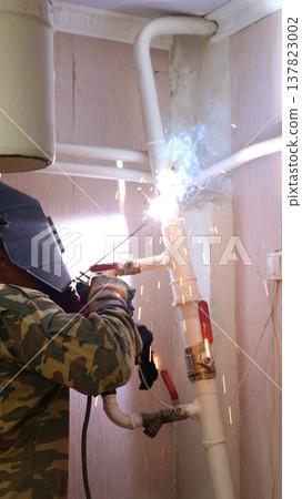 A welder, seen from behind, wearing a uniform and hard hat, welds a section of a house's heating system, made of white-painted iron pipes, to the ceiling. 137823002