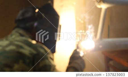 A welder, seen from the back, wearing a uniform and protective helmet, welds a house heating radiator made from white-painted iron pipes, a blurred image of an unrecognizable welder in the process A welder, seen from the back, wearing a uniform and protective helmet, welds a house heating radiator made from white-painted iron pipes, a blurred image of an unrecognizable welder in the process 137823003