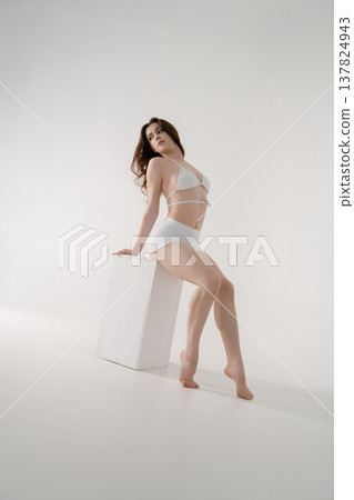 Graceful young woman in white lingerie posing on a cube in a minimalist high-key studio. 137824943