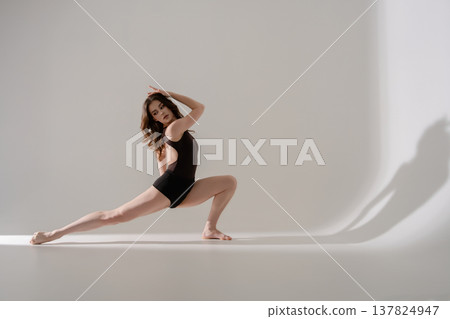 Graceful young woman in a black leotard performs a flexible lunge pose in a minimalist studio. Graceful young woman in a black leotard performs a flexible lunge pose in a minimalist studio. 137824947