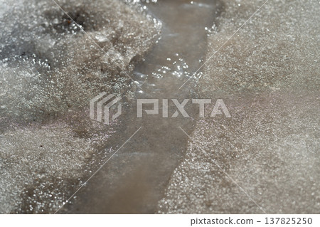 Spring flow over icy slush. Brown liquid and coarse gravel flow through melting icy slush. Muddy streams with gritty water trickle over melting ice in urban setting Spring flow over icy slush. Brown liquid and coarse gravel flow through melting icy slush. Muddy streams with gritty water trickle over melting ice in urban setting 137825250
