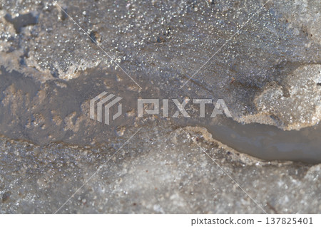 Flowing dirty snow melts along curb. Melted ice reveals siltladen water carving pathways. Environmental thaw exposes grooves in melting ice through muddy snow and water Flowing dirty snow melts along curb. Melted ice reveals siltladen water carving pathways. Environmental thaw exposes grooves in melting ice through muddy snow and water 137825401