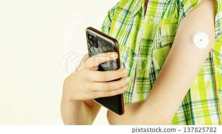 Teen holding smartphone with glucose sensor on arm against light background 137825782