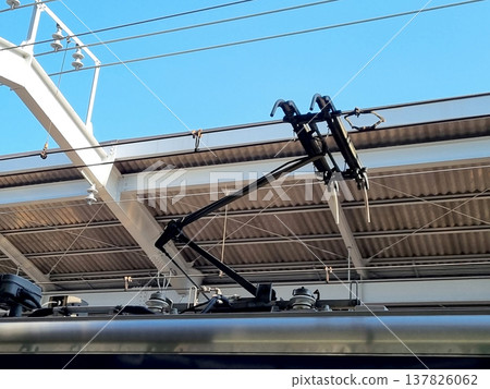 Close up of an electric train pantograph connected to overhead power lines at a railway station. Industrial details of public transportation infrastructure against a clear blue sky background. Close up of an electric train pantograph connected to overhead power lines at a railway station. Industrial details of public transportation infrastructure against a clear blue sky background. 137826062