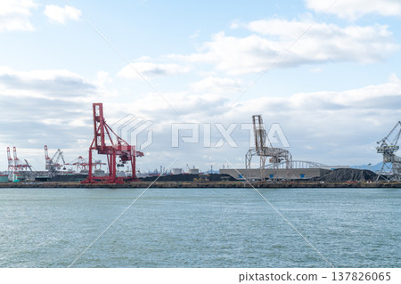 Industrial harbor with red gantry cranes unloading cargo near large piles of coal. Shipping port infrastructure and heavy machinery under a bright cloudy sky with blue water in foreground. Industrial harbor with red gantry cranes unloading cargo near large piles of coal. Shipping port infrastructure and heavy machinery under a bright cloudy sky with blue water in foreground. 137826065