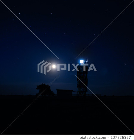 Silhouette of a navigation tower and a lonely tree against a deep blue night sky with stars and a bright moon. Peaceful maritime concept of safety, guidance, and hope in the dark. Silhouette of a navigation tower and a lonely tree against a deep blue night sky with stars and a bright moon. Peaceful maritime concept of safety, guidance, and hope in the dark. 137826557