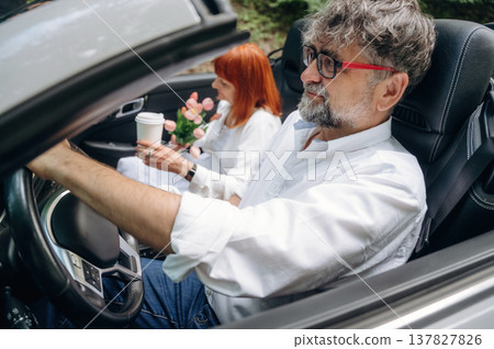 Cup of drink in woman's hand. Senior couple driving convertible cabriolet car Cup of drink in woman's hand. Senior couple driving convertible cabriolet car 137827826