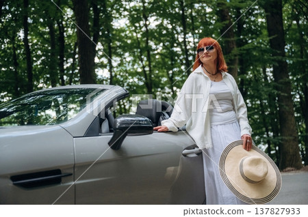 Woman with hat in hand is standing near cabriolet car on the road against trees Woman with hat in hand is standing near cabriolet car on the road against trees 137827933